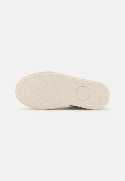 Såle av en sko laget av teksturert, lys beige gummimateriale, med et sirkulært detaljer og avrundet form.