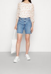 Blusa floreale con maniche trasparenti, abbinata a pantaloncini di jeans a vita alta con orlo sfrangiato. Sneakers bianche e una borsa a tracolla bianca completano il look.