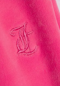 Šviesiai rožinės spalvos fliso drabužis su siuvinėta raide "J", demonstruojantis švelnią tekstūrą ir lygią apdailą.