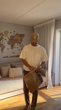 Brown parcel bag with black text, light-colored short-sleeve t-shirt, beige cap, and beige pants. Wooden world map on the wall.