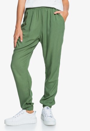 Femme portant un pantalon jogger vert ample avec taille et poignets élastiques, associé à des baskets blanches et un haut blanc.