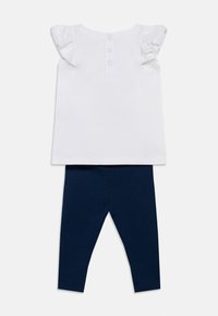 Haut blanc avec manches à volants, fermeture boutonnée au dos. Leggings bleu marine, coupe ajustée, confectionnés en matière douce.