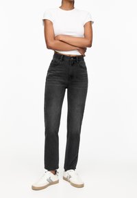 Jeans de mezclilla negros de talle alto con corte ajustado, cinco bolsillos y cierre de botón, combinados con zapatillas blancas y una camiseta blanca ajustada.