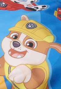 Personaggio dei cartoni animati con pelliccia marrone, cappello giallo e camicia blu, sorridente e che indica. Lo sfondo è di un azzurro chiaro con altri personaggi.