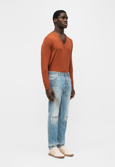 Homme debout portant un pull orange à manches longues avec col en V, un jean déchiré bleu clair et des baskets beiges, devant un fond uni.