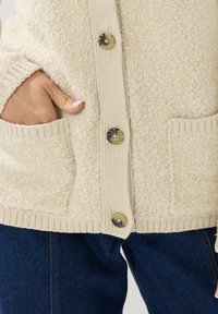 Maglione color crema con un tessuto morbido e texture. Presenta grandi bottoni, polsini a costine e tasche a toppa frontali.