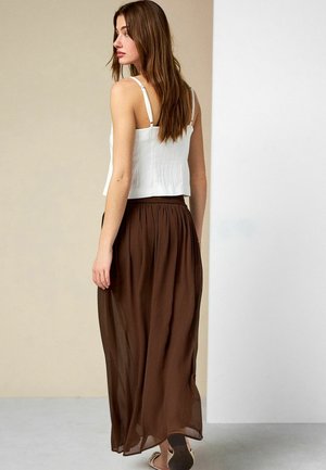 Frau mit langem braunem Haar, die ein weißes ärmelloses Oberteil und einen fließenden braunen Maxirock trägt, seitlich vor einem neutralen Hintergrund stehend.