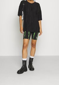 Top oversize plissettato nero, pantaloni bike tie-dye neri e verdi, ankle boots in pelle nera con suole spesse, calzini bianchi e una borsa a righe.