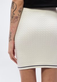 Λευκή πλεκτή φούστα με σχέδιο καλωδίου, υφασμάτινη υφή, εφαρμοστή γραμμή και ριγέ γιακά σε ναυτικό μπλε στο τελείωμα. Γυμνό χέρι με ορατά τατουάζ.