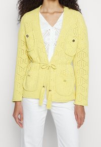 Żółty, dziergany kardigan z teksturowanym wzorem, wiązany w talii, z dwoma przednimi kieszeniami zapinanymi na okrągłe guziki oraz zdobieniami w postaci frędzli przy wiązaniu.
