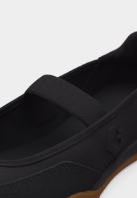 Chaussure de style Mary Jane noire avec bride élastique, tige en toile et détail de semelle gomme texturée sur fond blanc.