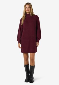Vestido de malha burgundy com gola alta, mangas longas e detalhes canelados na barra e nos punhos, combinado com botas pretas de cano alto.