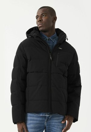 Chaqueta de plumas negra con capucha, diseño acolchado, cierre de cremallera, bolsillo en el pecho y puños elásticos. Llevada sobre una camisa de mezclilla, tejido texturizado.
