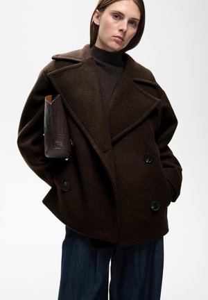 Vrouw draagt een donkerbruine oversized wollen jas met grote revers, zwarte knopen, een donker coltrui, blauwe broek, en houdt een bruine gestructureerde clutch vast.