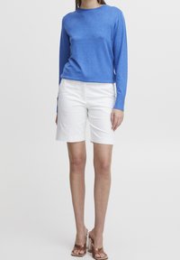 Femme portant une chemise à manches longues bleue, un short blanc à hauteur des genoux et des sandales à talons blocs transparents, debout devant un fond blanc uni.