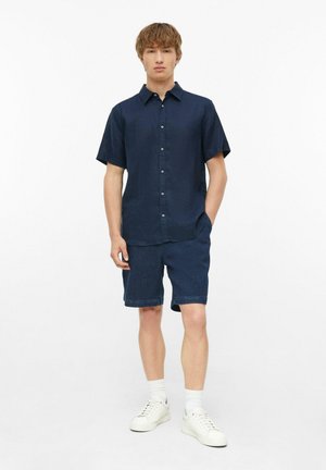 Jonge man staand, gekleed in een marineblauw kort mouw knoopoverhemd, marineblauwe shorts, witte sokken en witte sneakers op een witte achtergrond.
