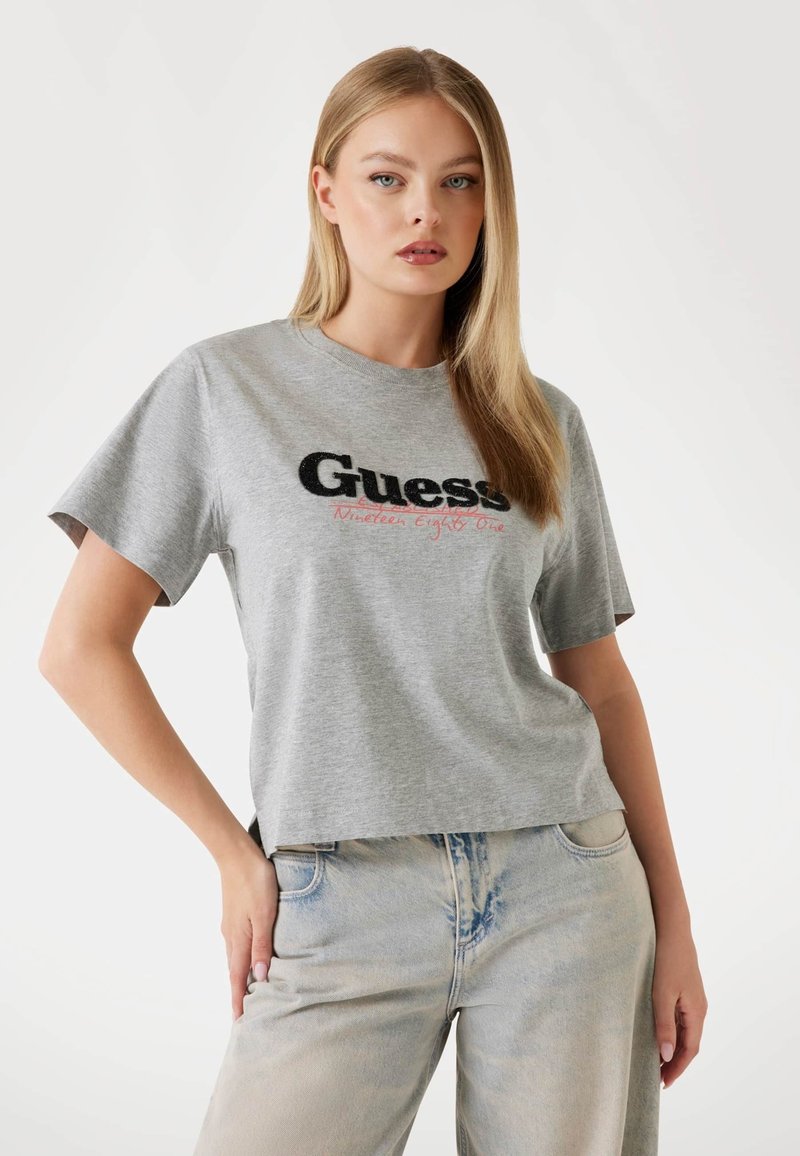 Graues kurzärmliges T-Shirt mit schwarzem und rotem "Guess"-Logo. Weiche Textur; lockere Passform. Kombiniert mit ausgebleichten blauen weit geschnittenen Jeans.