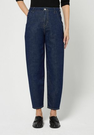 Frau trägt dunkelblaue Jeans mit geradem Bein und schwarze Slip-on-Schuhe vor schlichtem hellem Hintergrund.