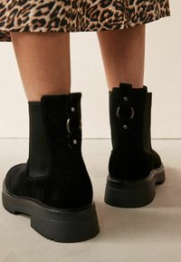 Mustad mustad ankle boots paksu taldaga, kaunistava rihmaga ja metallist detailide kujundusega taga. Tekstuuriga viimistlus, minimalistlik disain.