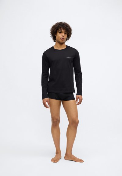 Top preto de manga longa com decote redondo e detalhe sutil do logótipo, combinado com calções pretos ajustados. Modelo está descalço sobre um fundo simples.