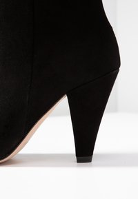 Bottine à talon haut en daim noir avec un talon effilé et une semelle beige sur une surface blanche et un fond clair.