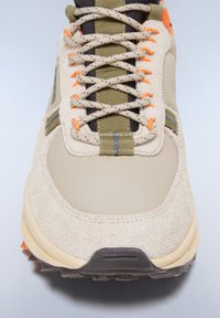 Světlé semišové tenisky s béžovými akcenty, oranžovými tkaničkami a zelenými textilními detaily. Mají strukturovanou podrážku a kontrastní kovové detaily.