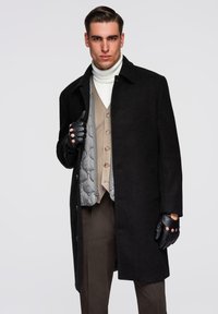 Schwarzer Wollmantel mit spitzem Kragen, darüber getragen eine hellgraue, gesteppte Weste und ein beigefarbenes Hemd mit Knöpfen. Schwarze Lederhandschuhe mit ausgeschnittenen Details.