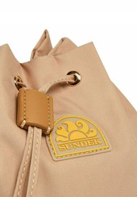 Borsa a drawstring beige realizzata in tessuto morbido con una texture opaca, caratterizzata da un logo del brand in gomma gialla e hardware in metallo color oro.