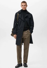 Schwarzer Trenchcoat mit großem Kragen, doppelt-knöpfig, kombiniert mit einem schwarzen Rollkragenpullover und khakifarbenen Hosen. Er trägt schwarze Schuhe und eine Tasche.