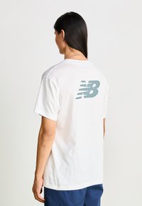 Bílá bavlněná tričko s krátkým rukávem, na zádech s velkým světle modrým logem New Balance. Hladká textura a uvolněný střih.