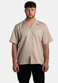 Chemise à manches courtes kaki avec un design à boutons, texture lisse et logo brodé sur le devant à gauche. Portée avec un pantalon noir.