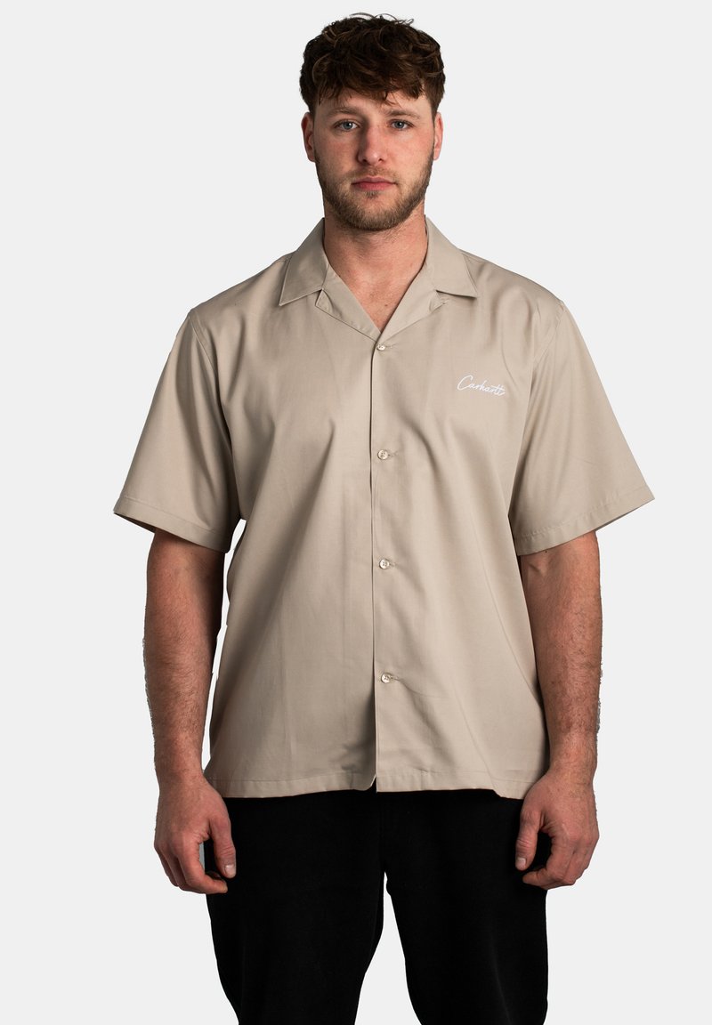 Camisa de manga corta en color caqui con un diseño de botones, textura suave y un logotipo bordado en el lado izquierdo delantero. Se lleva con pantalones negros.