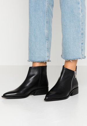 Zwarte leren enkellaarzen met zijrits, gedragen met lichtblauwe cropped jeans met rafelige zoom, tegen een effen witte achtergrond.