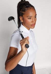 Polo bianco con tre bottoni e dettagli neri. Tiene una mazza da golf, capelli intrecciati. Pantaloni blu navy e tatuaggio dettagliato sul braccio.