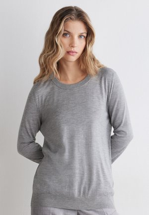 Ung kvinde med bølget blondt hår iført en lysegrå langærmet sweater, stående mod en ensfarvet hvid baggrund.