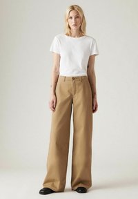 Crème-kleurig t-shirt met korte mouwen, gecombineerd met wijde, beige broeken. Beide items hebben een gladde, zachte textuur. Zwarte schoenen maken de look compleet.