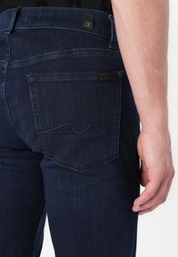Tamno plavi traperici s uskim krojem, dizajnom stražnjeg džepa sa suptilnim šavovima i teksturiranom završnom obradom. Oznaka branda vidljiva na pojasu.