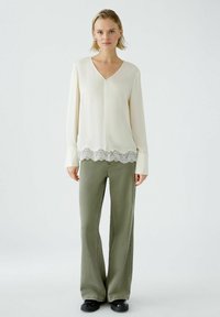 Weißes Langarmbluse mit V-Ausschnitt und Spitzenabschluss, kombiniert mit olivgrünen Schlaghosen. Schwarze Schuhe runden das Outfit ab.