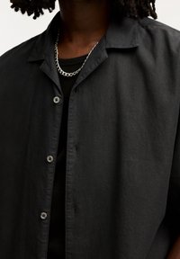 Camicia nera a maniche corte realizzata in tessuto leggero, caratterizzata da un colletto e chiusura a bottoni, indossata sopra un top aderente nero.