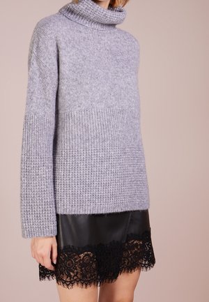 Vrouw draagt een lichtgrijze, gebreide coltrui met een gestructureerd patroon en een zwarte leren rok met zwarte kanten rand.