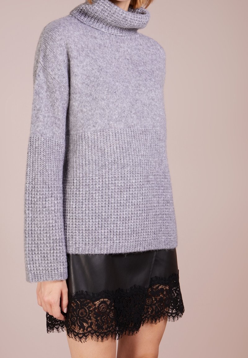 Femme portant un pull à col roulé en maille gris clair avec un motif texturé et une jupe en cuir noir ornée d'une bordure en dentelle noire.