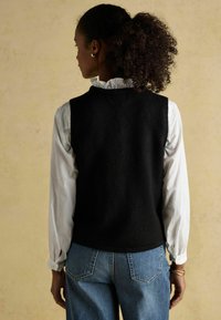 Mustade villaste väled üleval valge volangiga särk, koos siniste teksadega. Vestil on puhas, struktureeritud kuju ja pehme tekstuur.