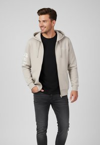 Beige Zip-up-Hoodie mit einer Fronttasche, Kapuze mit Kordelzug und weißem Text auf dem Ärmel. Schwarzes T-Shirt darunter, kombiniert mit grauen Jeans.