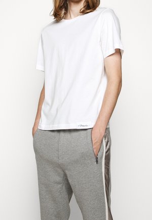 Person iført en ensfarvet hvid kortærmet t-shirt og grå joggingbukser med sidelommer med lynlås og skinnende striber, hænder i lommerne.