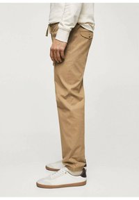 Pantalon en coton beige à coupe ajustée, avec poches latérales et taille à cordon. Porté avec des baskets blanches et un sweat-shirt de couleur claire.