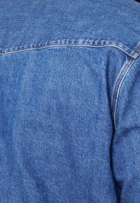 Jeansjacka i en medelblå nyans. Den har synlig sömnad, en standardkrage och en texturerad tygytan med lätt blekning.