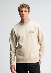 Beige sweatshirt met ribgebreide manchetten en zoom, met een kleine zwarte logo-print op de linkerkant van de borst. Gladde textuur, ronde hals.