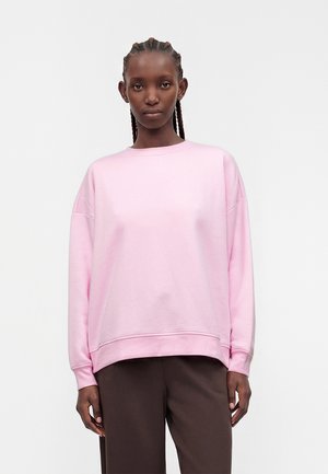 Jong persoon met vlechten, gekleed in een lichtroze longsleeve trui en donkerbruine broek, staand tegen een effen achtergrond.