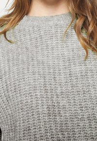 Pull en maille grise avec un motif texturé, doté d'un col rond et de manches longues. Tissu épais et doux avec des coutures visibles.