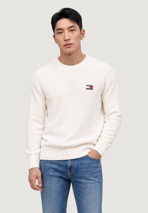 Hombre con un suéter de punto blanco con un pequeño logo rojo y azul en el pecho y vaqueros azules, de pie con una mano en el bolsillo.